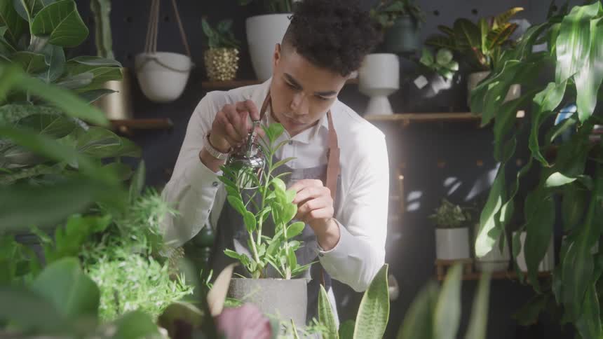 Man uses a metal container to spray a plant while standing among shelves of plants
