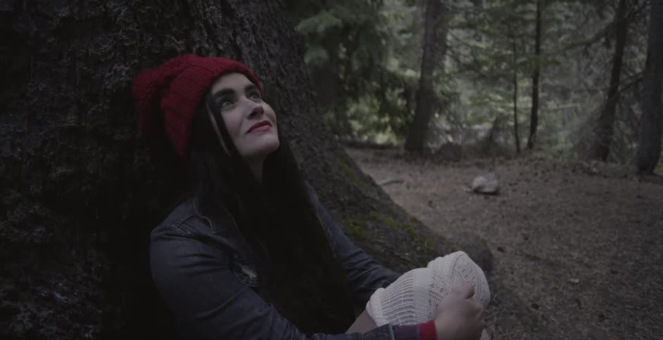 A woman is sitting at the base of a tree and looking around at a forest