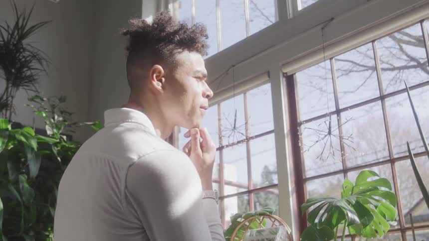 A man standing with his hand on his chin looking out a window in an interior with bright natural light coming from outside