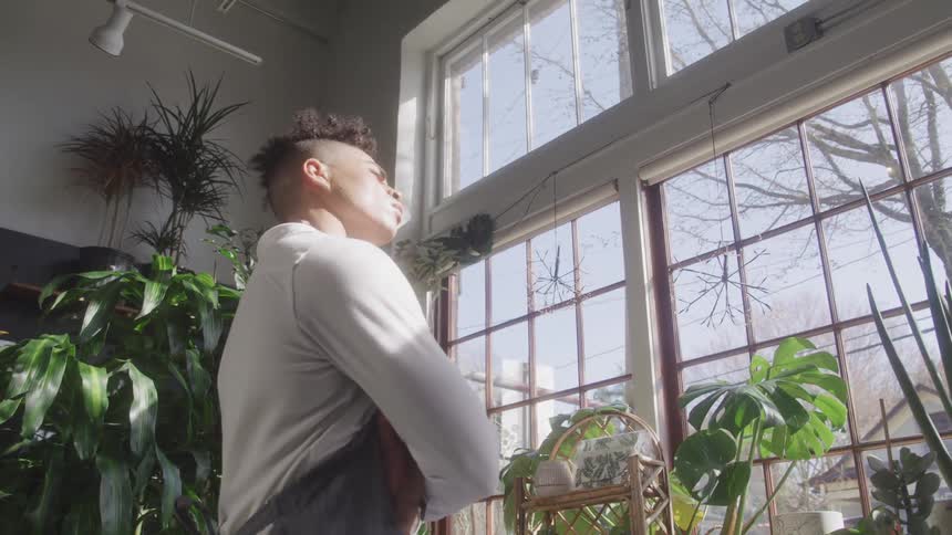 A man stands in a room full of potted plants and looks out a window
