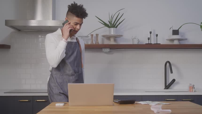 A man wearing a pair of panties is already walking in a kitchen talking on his cell phone and in front of him is a table with an open laptop