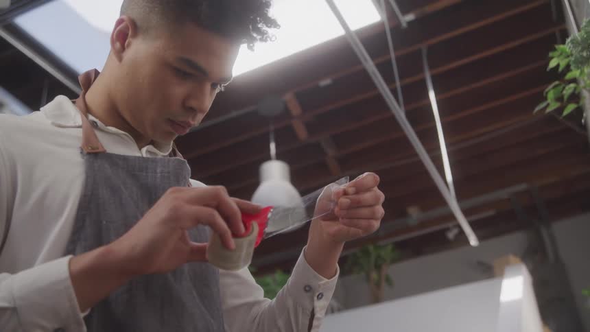 A young man stands in a green house while pulling tape from a dispenser
