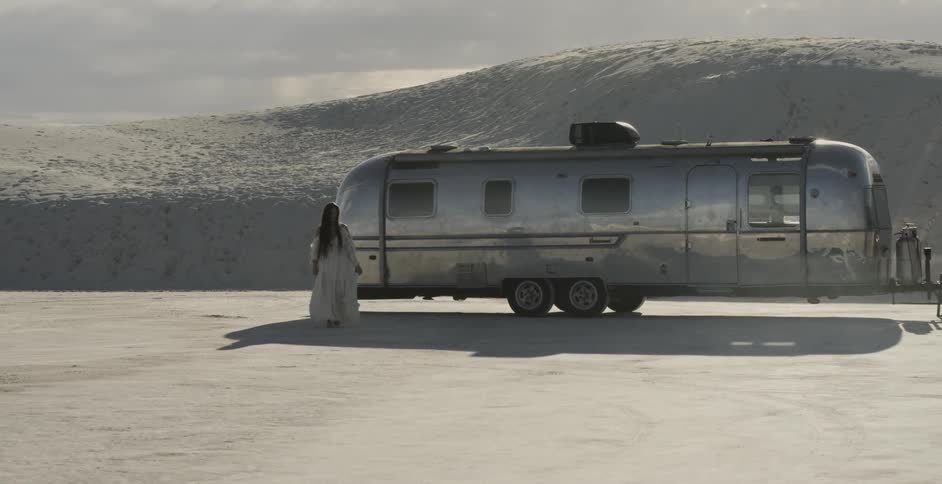Woman in white dress walking away from travel trailer in desolate region