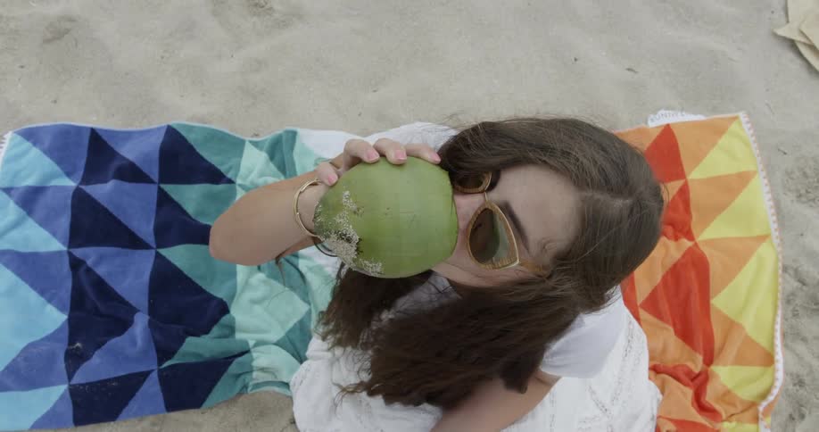 Woman sitting on towel in the sand drinking from coconut
