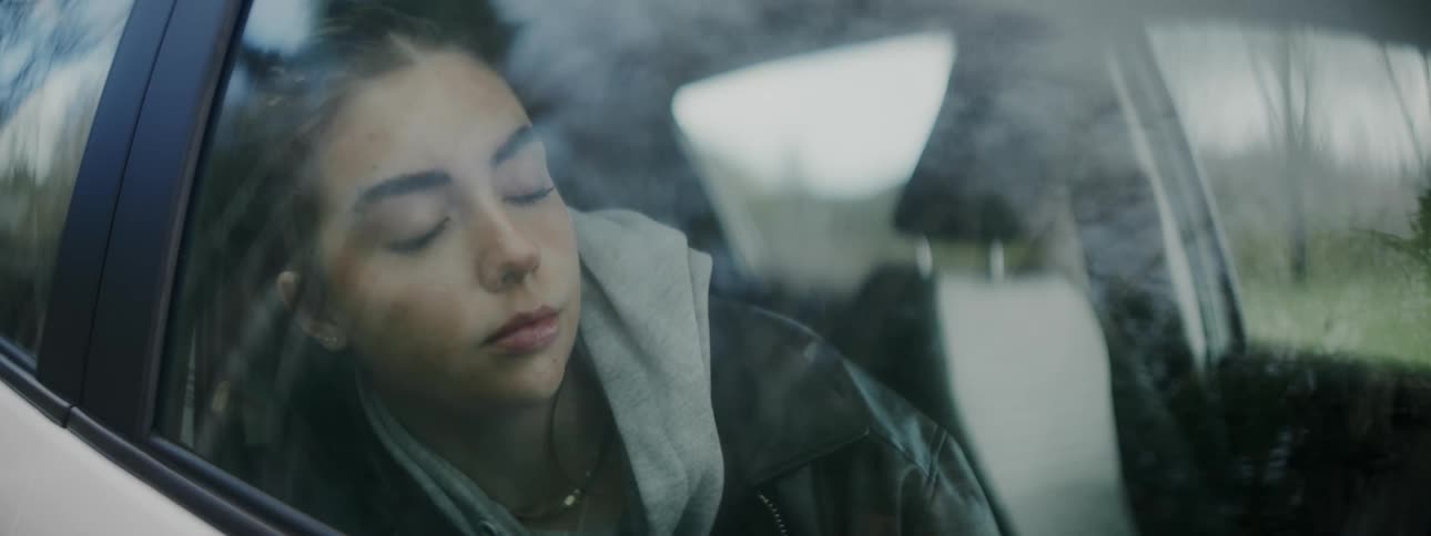 A teen girl wearing a hoodie is sleeping in a car with her head leaning against the passenger window