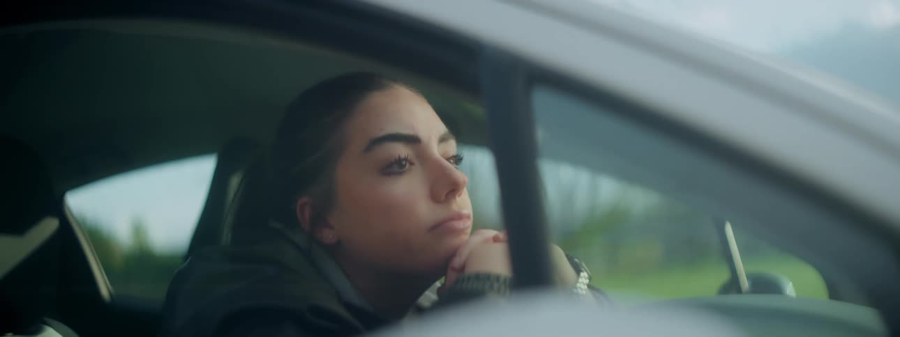 A woman sits in a parked car with her head resting on her hands which are placed on the steering wheel