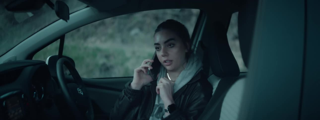 A woman wearing a hoodie under a jacket is seated in a cars drivers seat holding a phone to her ear while engaged in conversation with an outdoor background visible through the window