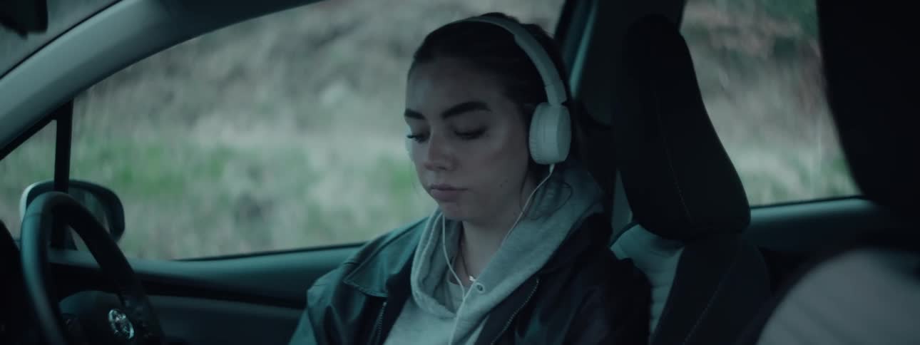 A young woman is sitting alone in a car and singing along to music shes listening to on headphones