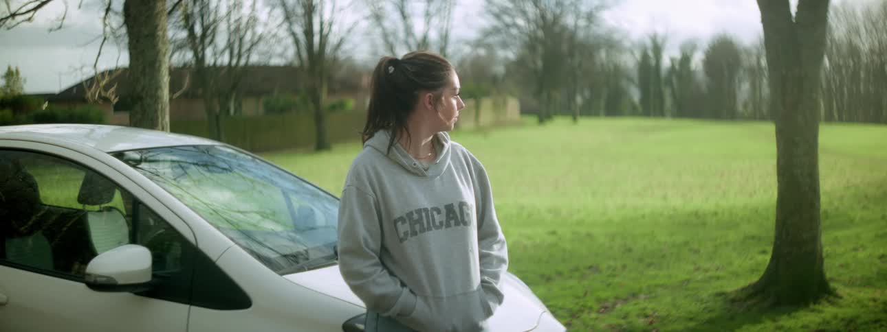 A woman in a gray sweater stands besides a car parked near a park