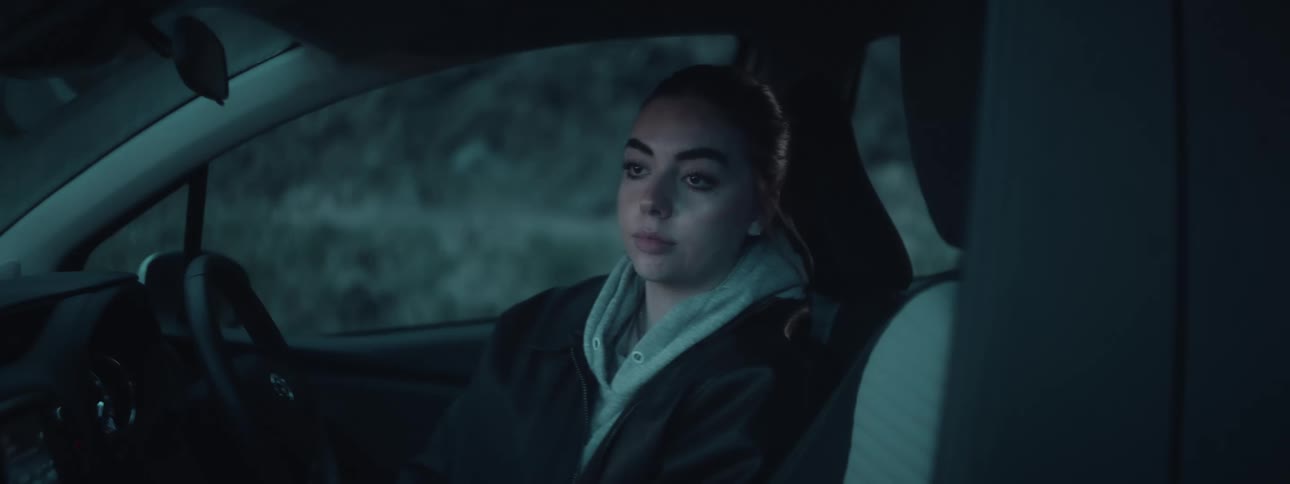 A woman wearing a hoodie under a jacket is seated in a cars drivers seat with an outdoor background visible through the window
