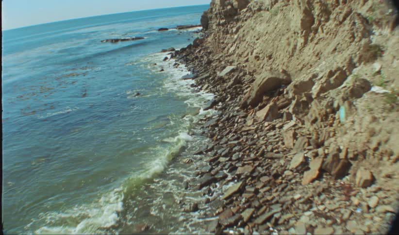 Waves crash against the rock strewn shore at the base of a cliff during the day