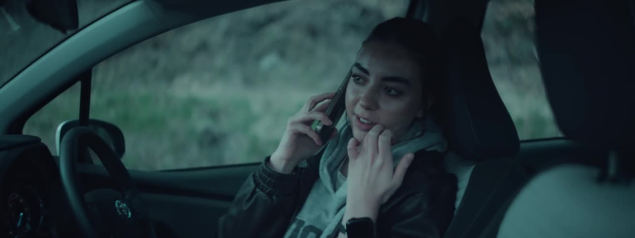 A young woman sits in the passenger seat of a car wearing a hoodie and leather jacket as she talks on the phone with a contemplative expression in a dimly lit moody atmosphere