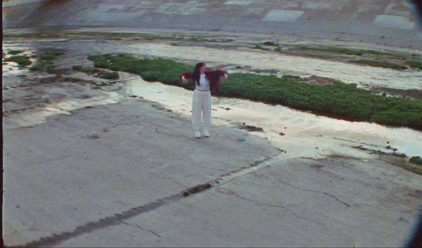 A woman in white pants and a red jacket dances on cracked concrete within a shallow drainage canal
