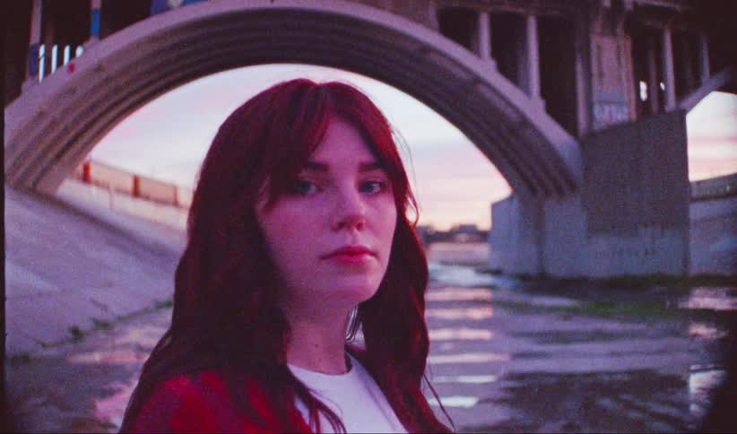 A woman stands near a concrete river channel under an arched bridge with the warm hues of sunset illuminating the scene