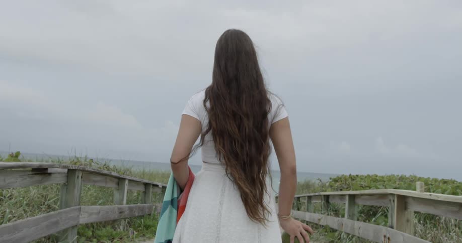 Woman holding towel and coconut standing on beach path near ocean
