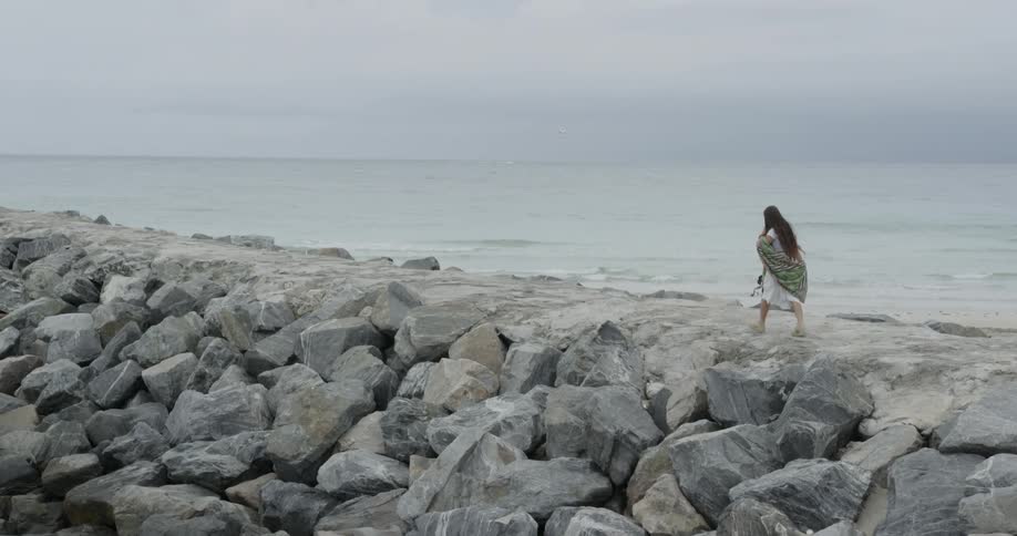 A woman walks across boulders beside the ocean