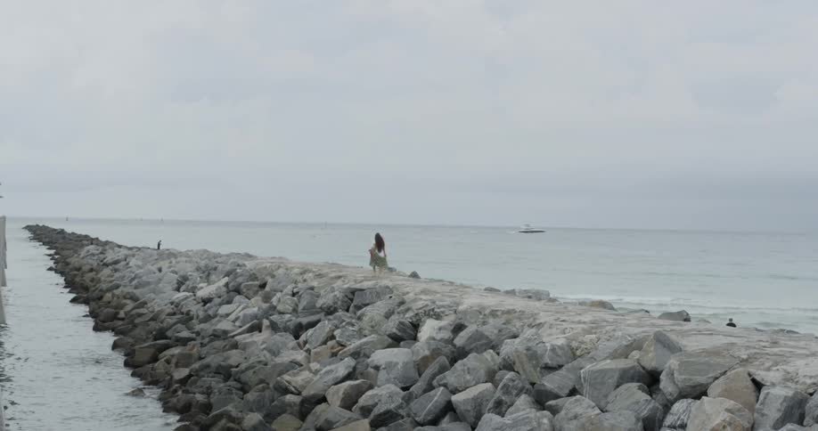 A woman is walking out on a long rock jetty on a cloudy day