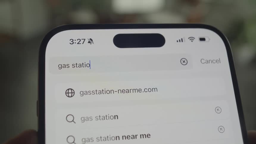 A closeup shot shows a smartphone screen displaying a search bar with the words gas stations near me and related search suggestions
