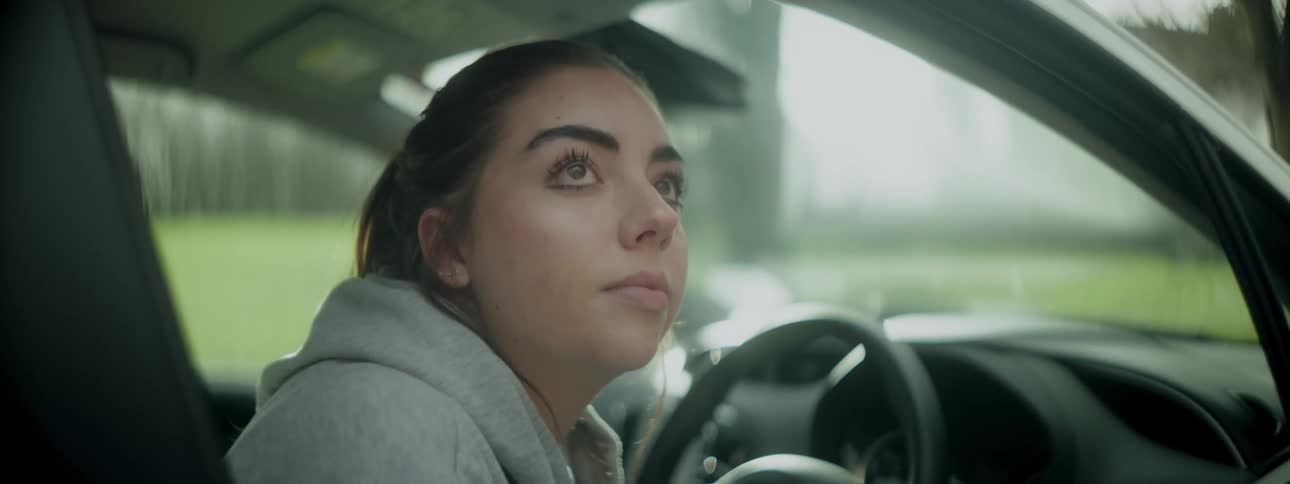 A woman wearing a hoodie under a jacket is seated in a cars drivers seat rolling down the window as she gazes outside