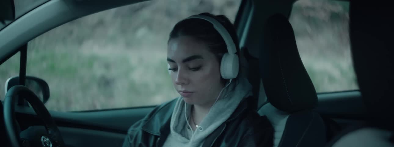 A woman wearing headphones and a hoodie sits in the drivers seat of a car