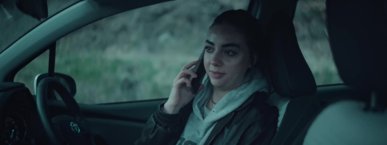 A woman wearing a hoodie under a jacket is seated in a cars drivers seat holding a phone to her ear while engaged in conversation with an outdoor background visible through the window