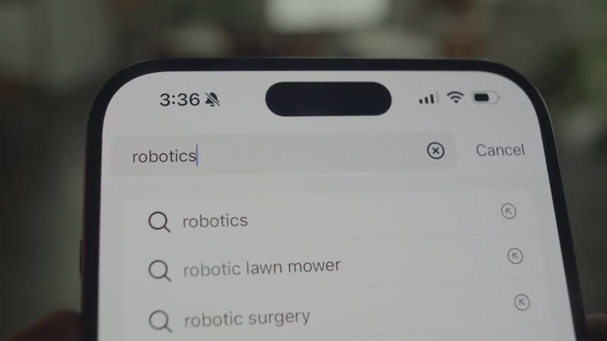 A closeup shot shows a smartphone screen displaying a search bar with the word robotics and related search suggestions