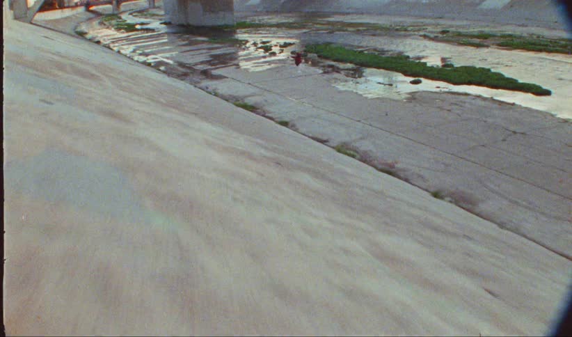 A woman in white pants and a red jacket dances on cracked concrete within a shallow drainage canal