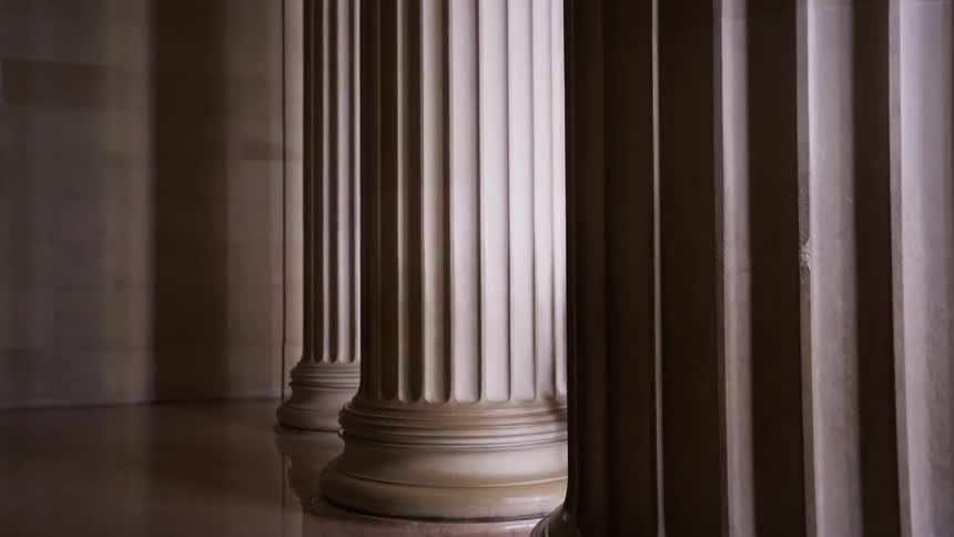A row of fluted classical columns stands on a reflective floor in a dimly lit interior