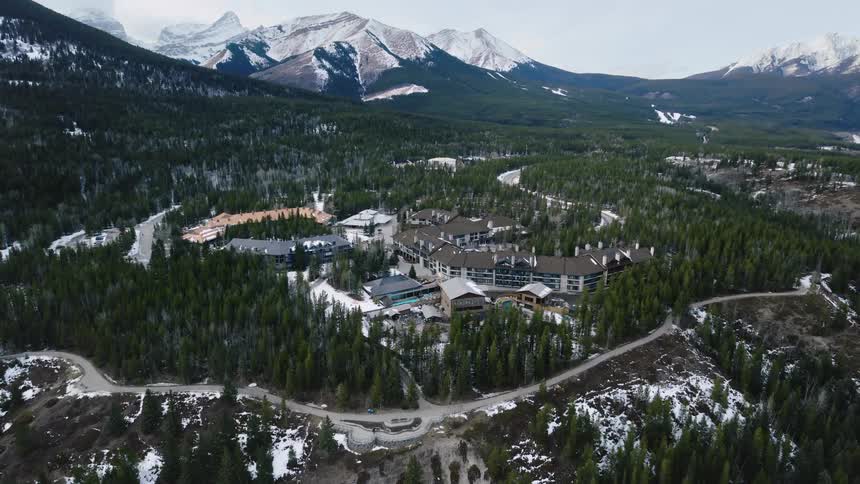 An aerial view captures buildings nestled amongst dense evergreen trees in a valley surrounded by snowdusted mountains under a gray sky