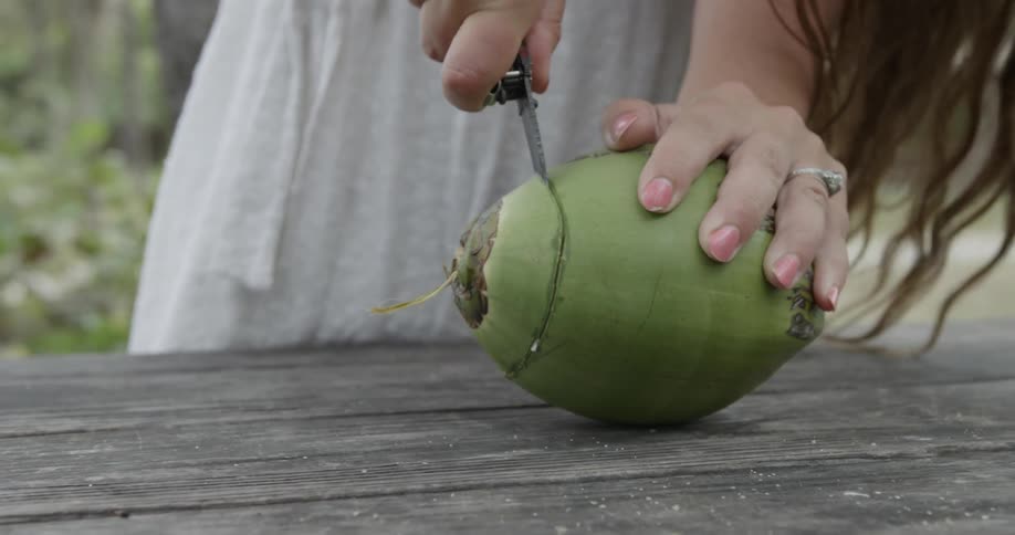 A woman is using a knife to cut a coconut on a picnic table