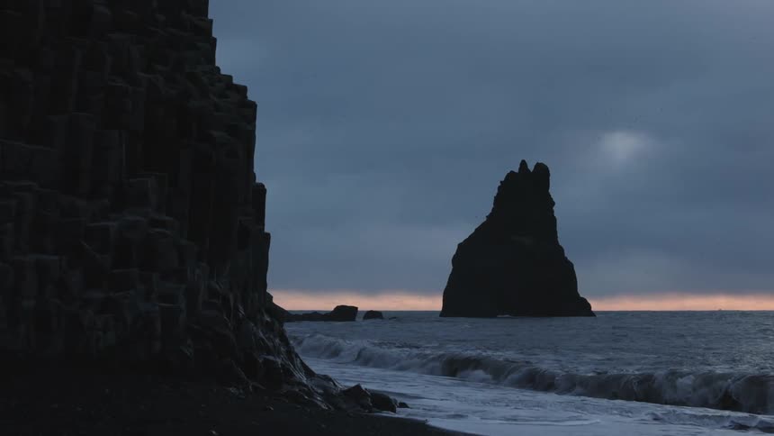 Near a towering basalt cliff a dark sea stack rises from the ocean under a moody sky with gentle waves lapping at the black sand shore and a subtle band of light glowing along the distant horizon in the fading twilight
