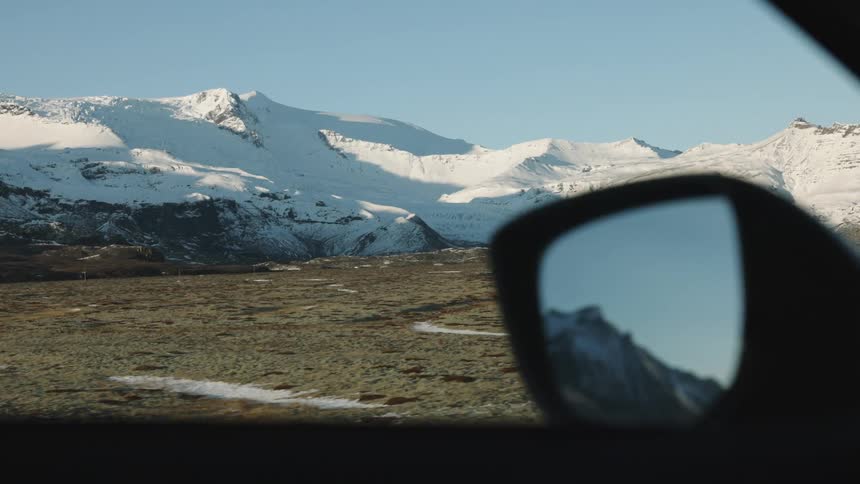 A cars side mirror frames a snowy mountain range against a clear sky viewed from inside the vehicle