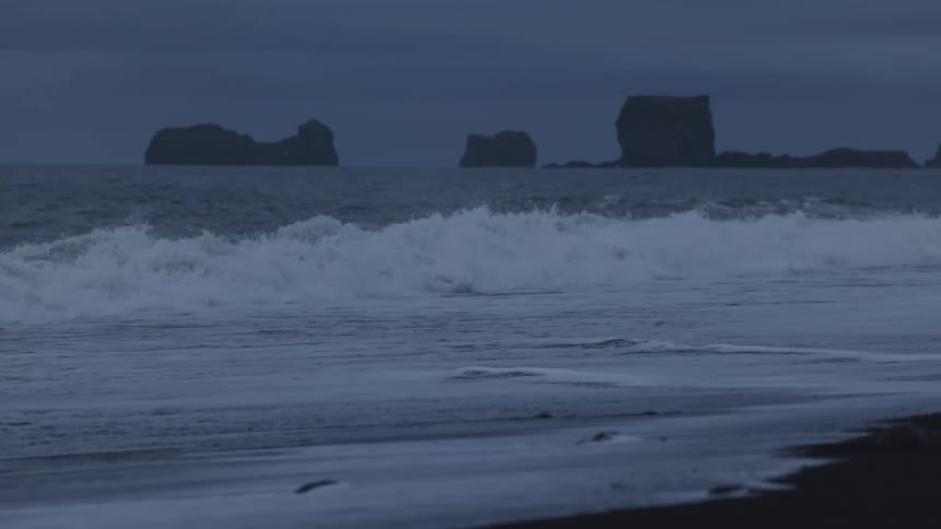 A dark moody seascape shows waves crashing on a black sand beach with silhouetted rock formations in the distance under an overcast sky
