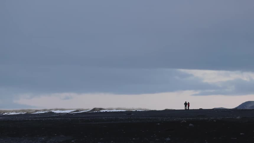 Two individuals stand in a vast open landscape beneath an overcast sky their figures outlined against the wide horizon and the dark ground with a subtle hint of snow visible near the distant edge