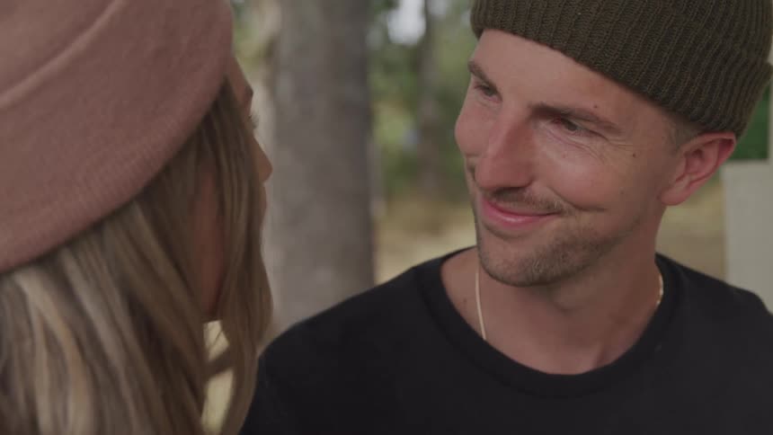A couple one in a pink beanie and the other in a green beanie sit outdoors talking to each other