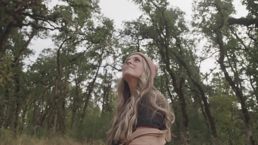 Woman with long blonde hair standing in middle of forest admiring the trees
