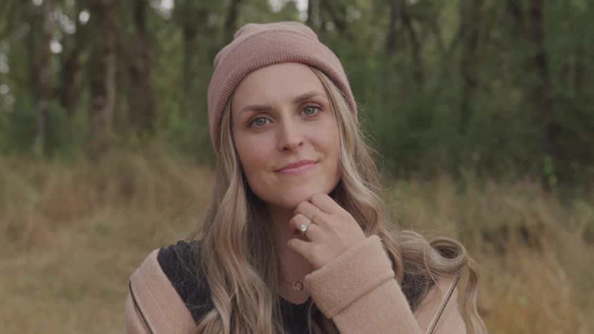 A woman with long wavy hair wearing a pink beanie and a beige jacket stands outdoors in a forested area holding a hand near her face
