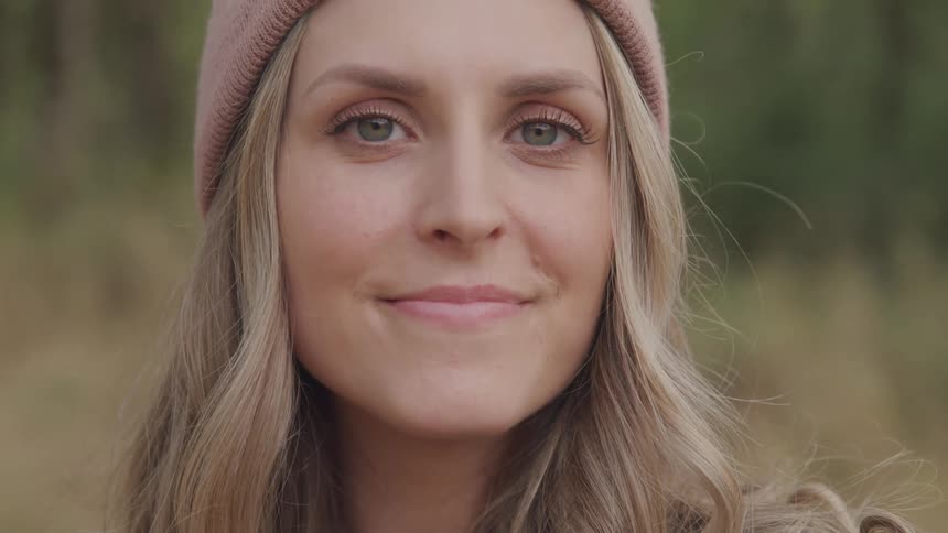 A closeup of a blonde woman wearing a beanie gazing directly at the camera in an outdoor setting