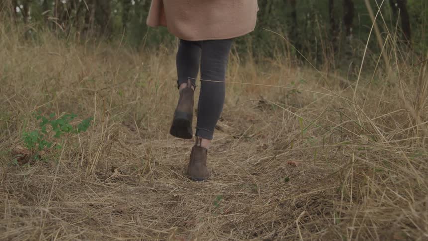 A woman wearing a beige coat dark leggings and brown ankle boots walks through a dry grassy field
