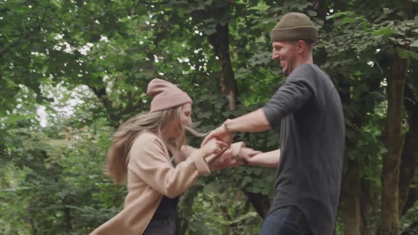 A couple are holding hands and running through a wooded area with trees and fallen leaves scattered on the ground