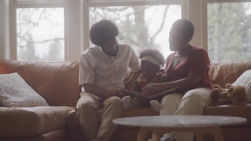 A family of three sits together on a brown couch in a living room reading a book with large windows and a round wooden coffee table in the foreground