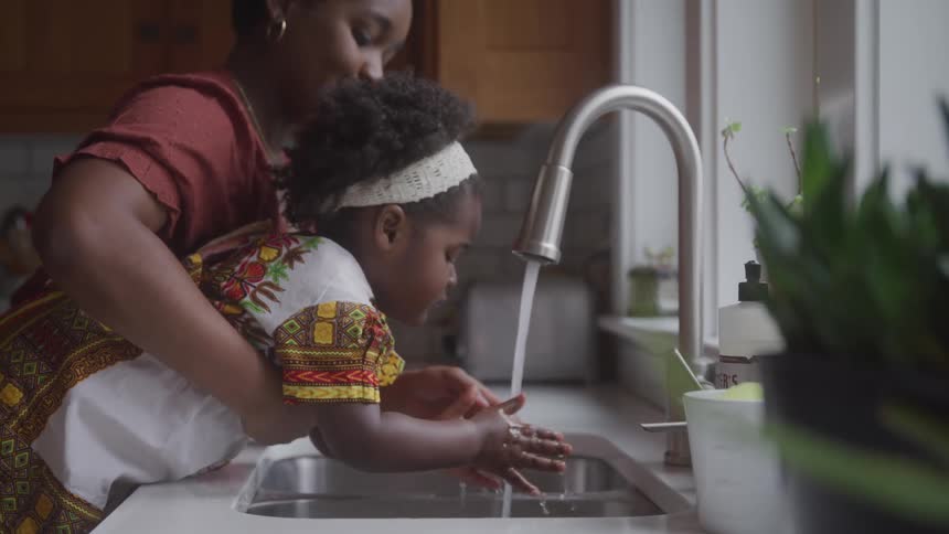 A little girl on the lap of a woman who is taking her to a sink with a tap with water to wash her hands