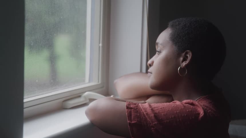A woman wearing a red shirt with textured patterns leans on a windowsill gazing outside through a window