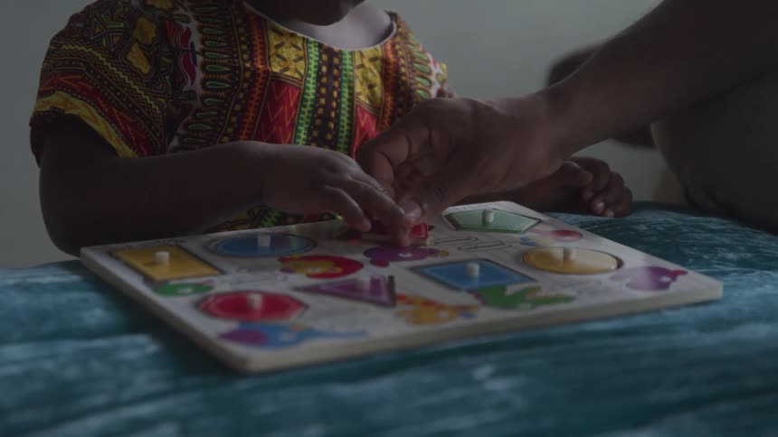 An adult assists a child in solving a colorful shape puzzle on a blue surface indoors