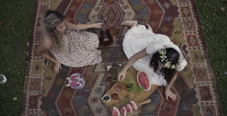 Two women sitting down on picnic blanket to eat watermelon