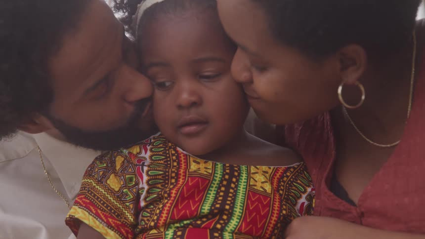 A closeup of a father and mother kissing their daughter who is dressed in a brightly colored patterned outfit with red yellow green and black designs