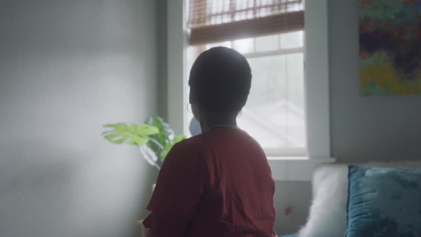 The back of a young woman sitting in the bed looking at a window on a sunny day