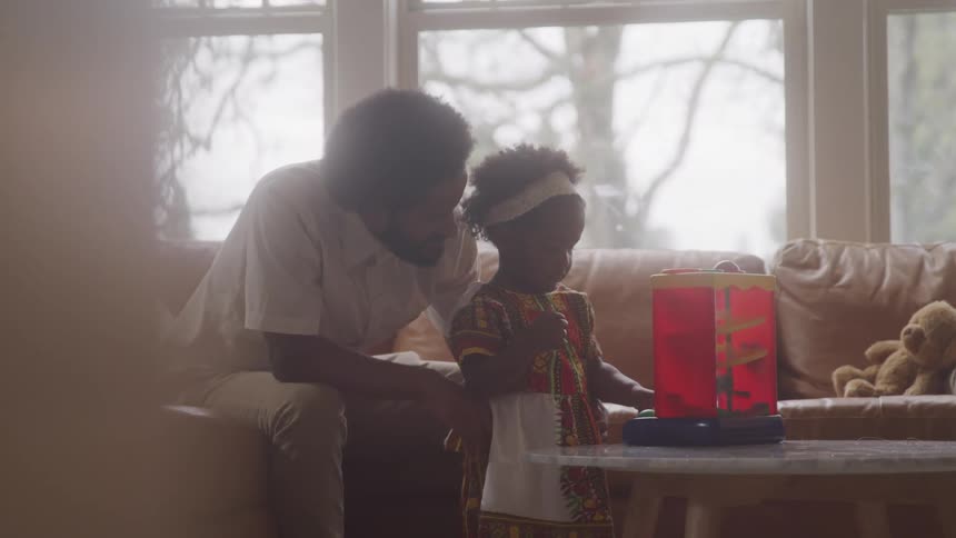 An adult man sitting on the sofa and looking at his daughter who is in front of him playing with a red object on top of a table