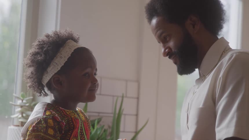 A bearded father gazes lovingly at his daughter who is wearing a white headband and a vibrant patterned outfit in a room near a window