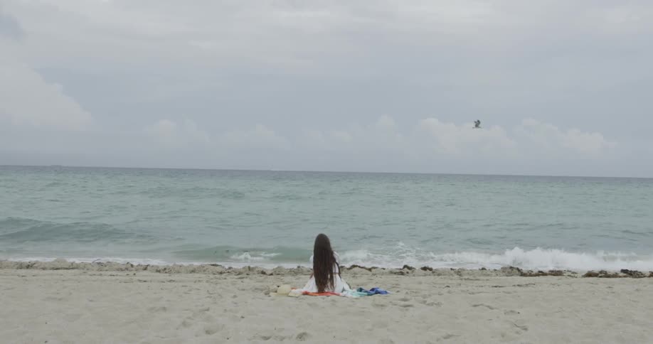 Woman in white dress sitting on blanket on the beach facing the ocean